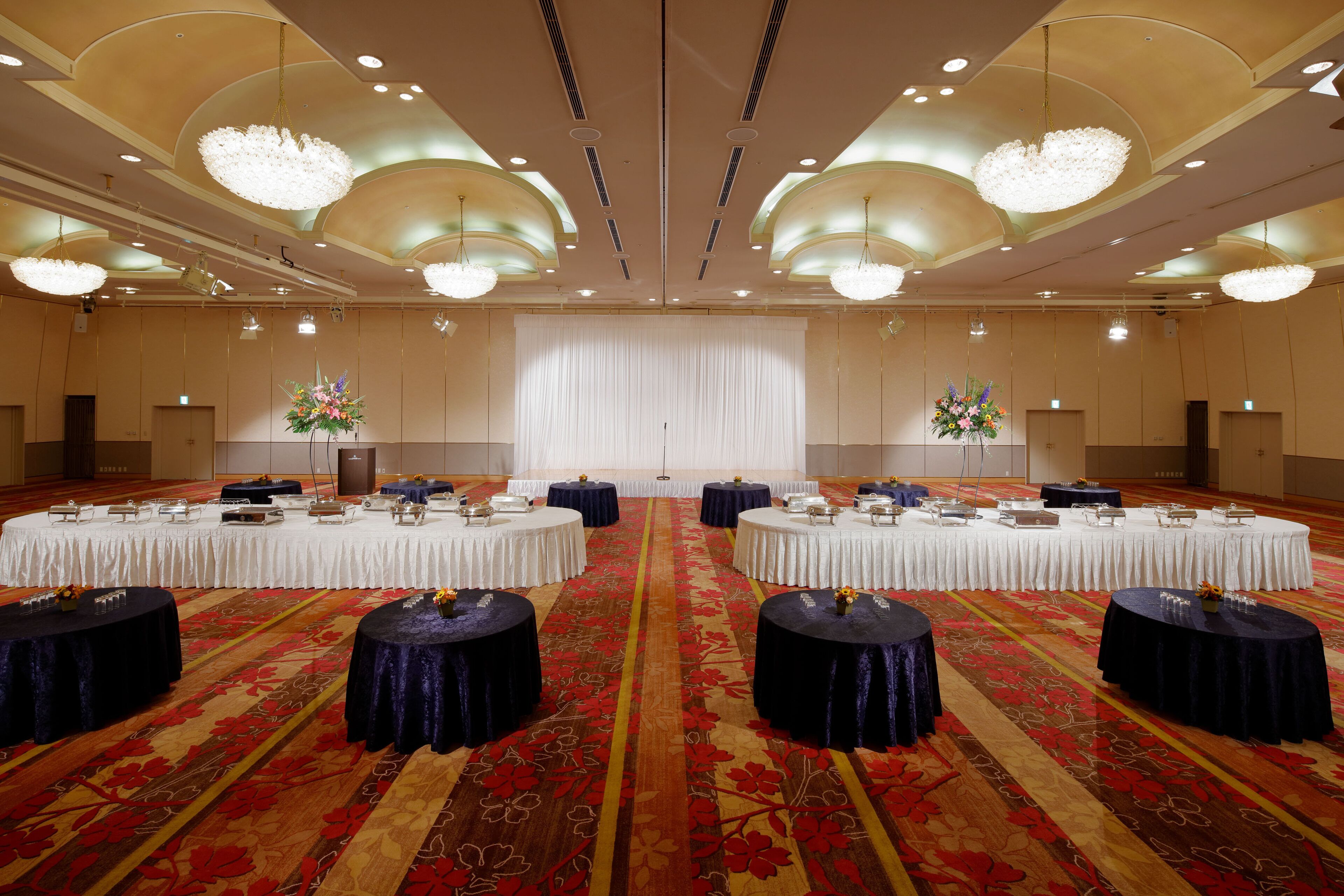 Un amplio salón de conferencias con mesas redondas, una alfombra estampada y candelabros.