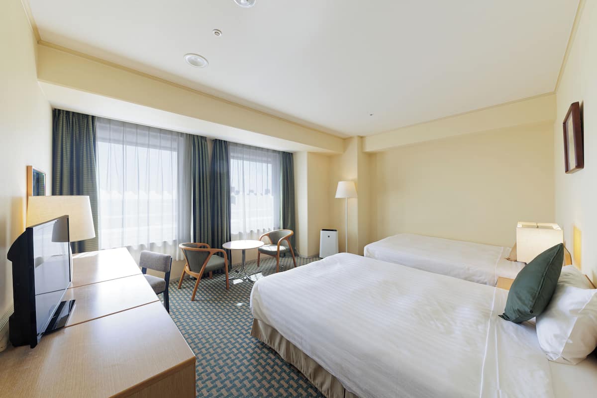 Una habitación de hotel con dos camas, un escritorio, una silla, una lámpara y una ventana con cortinas.