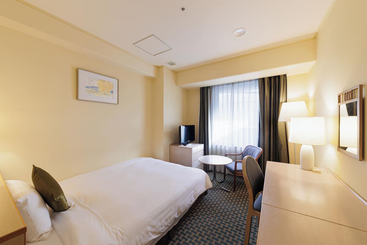 Habitación de hotel con cama, escritorio, silla, televisor, lámpara y ventana con cortinas.