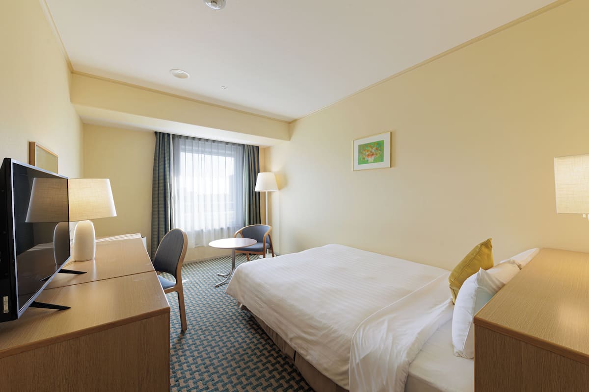 Habitación de hotel con cama, escritorio con computadora, silla, mesita y ventana con cortinas.