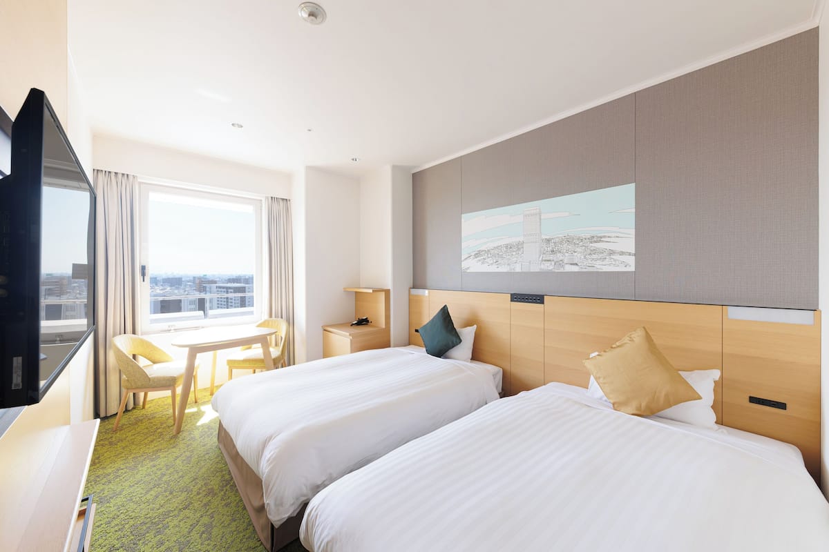 Una habitación de hotel con dos camas, un escritorio, una silla, un televisor y una ventana con vista a la ciudad.