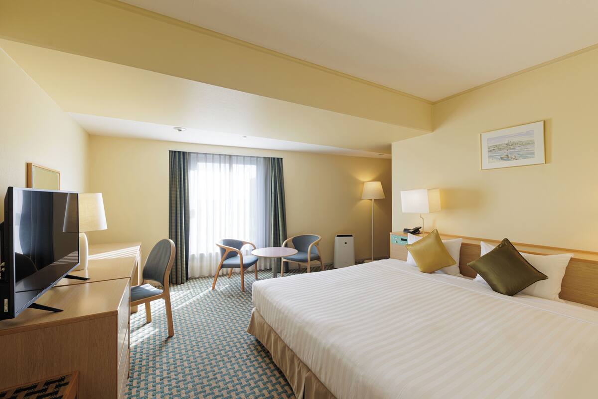 Habitación de hotel con una cama grande, un escritorio con televisor, dos sillas, una mesita y una ventana con cortinas.