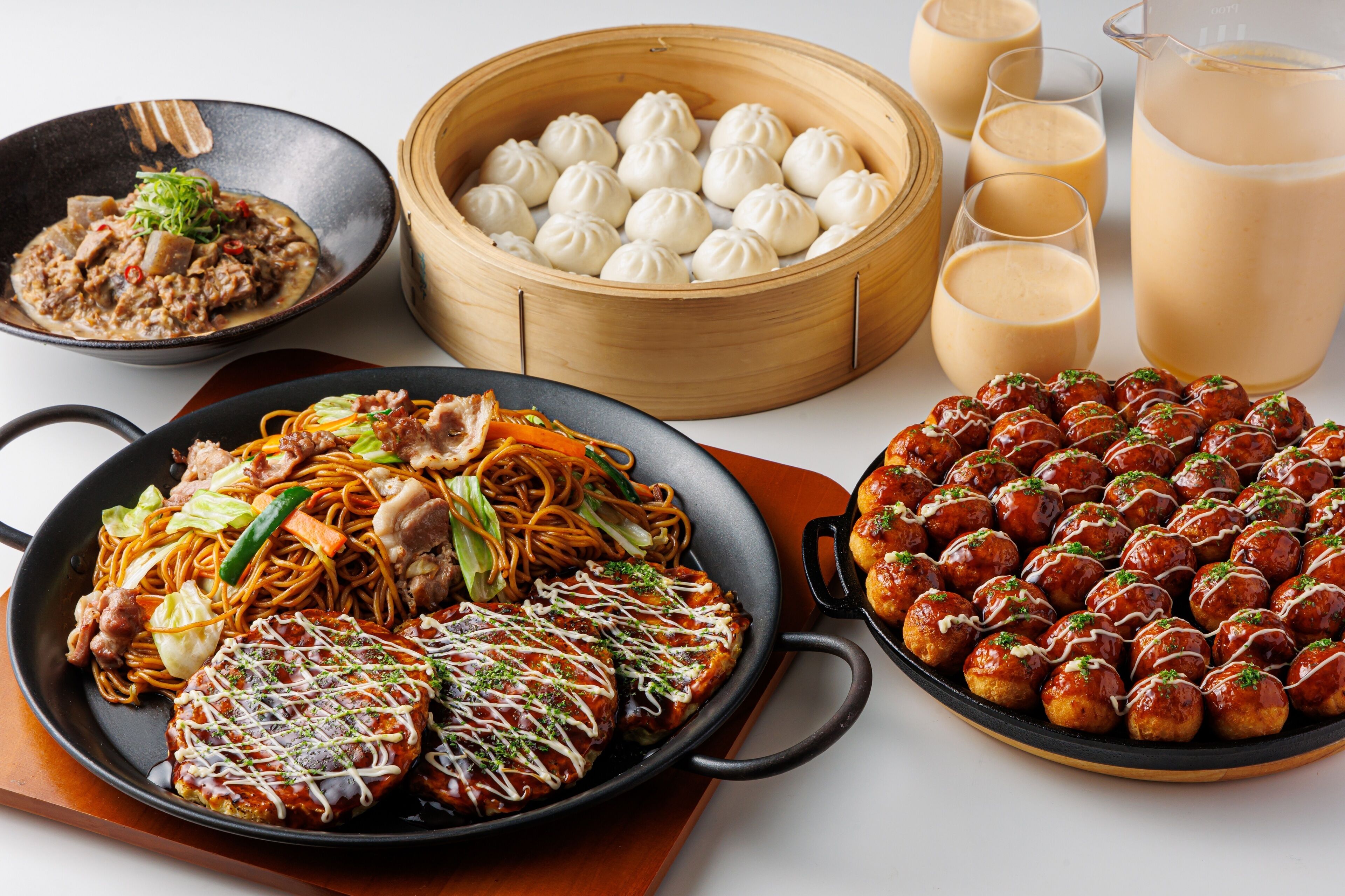 A variety of Asian dishes including dumplings, noodles, and meat dishes, along with several glasses of a light-colored beverage.