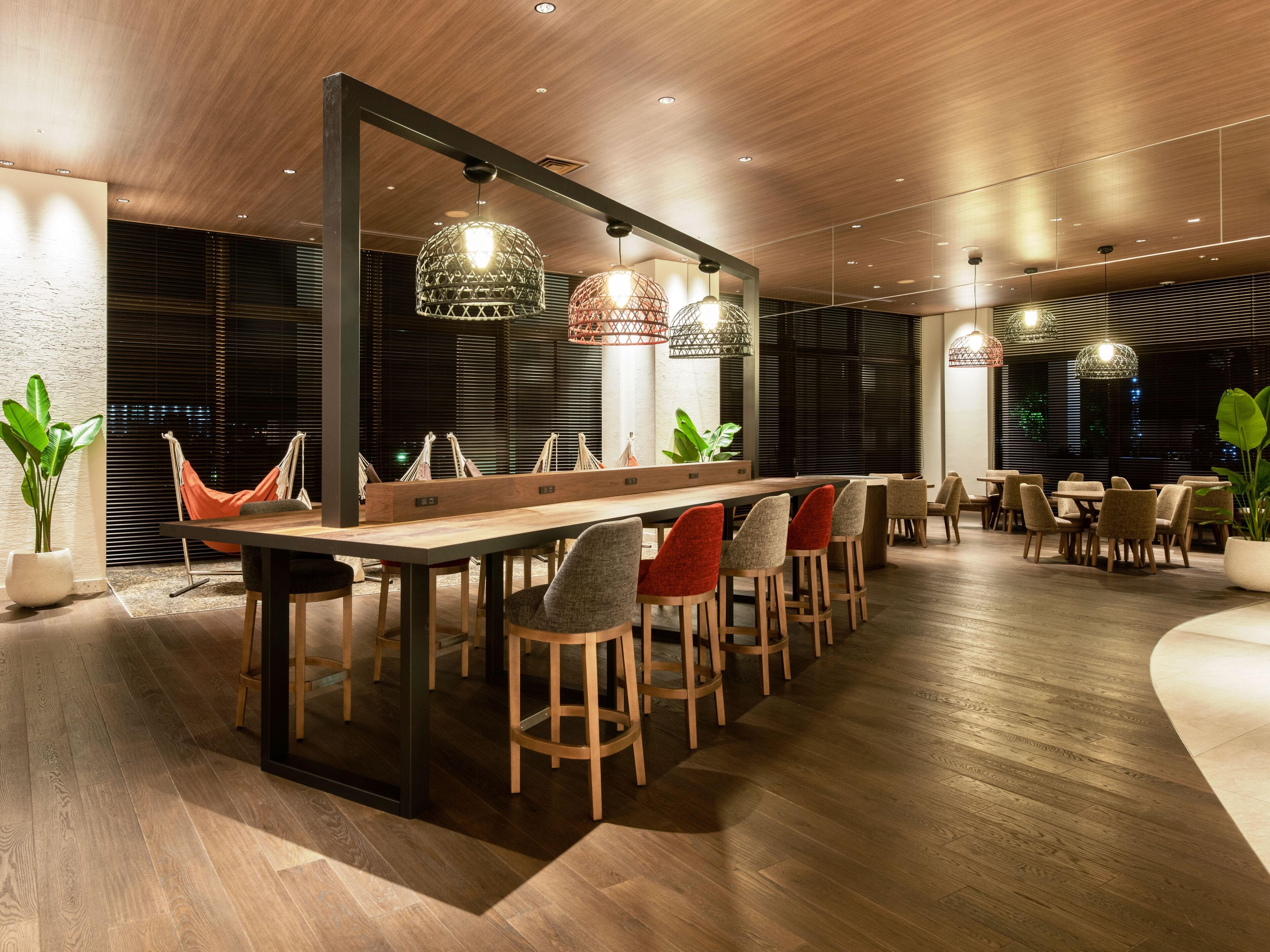 Un espace de salle à manger moderne, avec une longue table en bois, de nombreuses chaises et des luminaires suspendus.