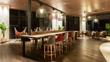 Un espace de salle à manger moderne, avec une longue table en bois, de nombreuses chaises et des luminaires suspendus.