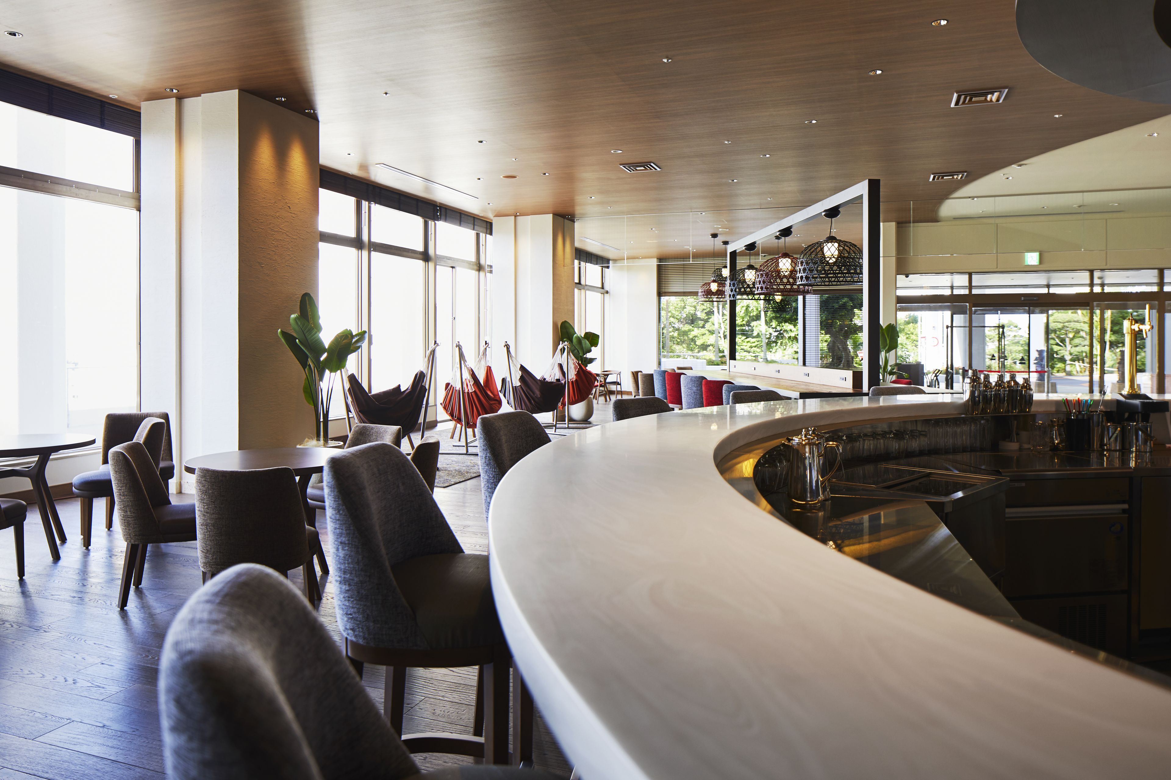 A modern bar area with a curved counter, seating with grey upholstered chairs, and large windows offering views of the outdoors.
