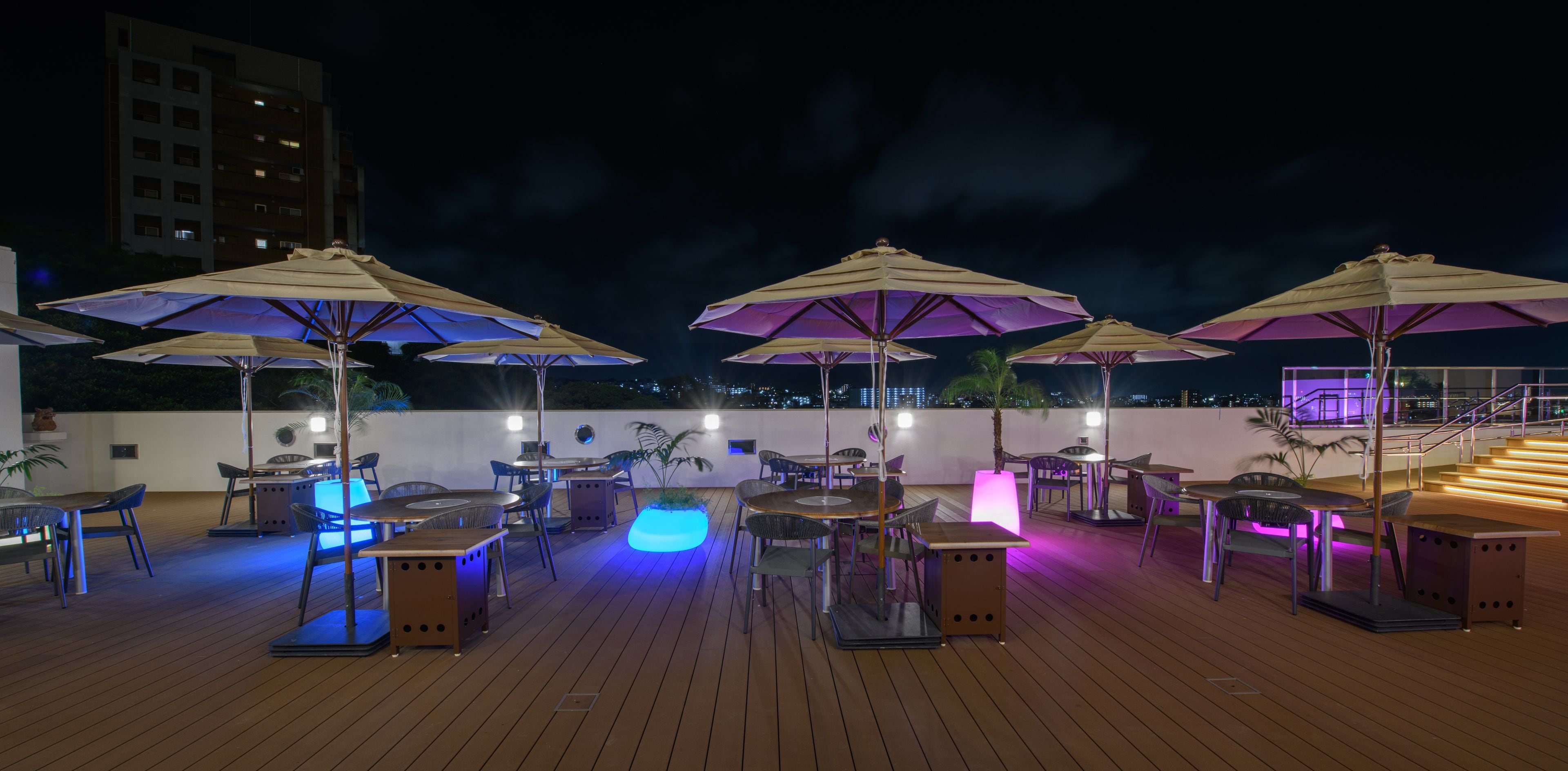 Terraza exterior con varias mesas y sillas, rodeada de sombrillas e iluminada con luz ambiental.
