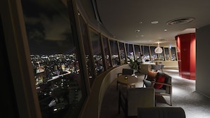 A modern hotel room with a curved balcony, large windows offering a city view, a dining area with a table and chairs, and a red accent wall. - INFINISIS NAHA SHURIJO by Hewitt Resort (Naha)
