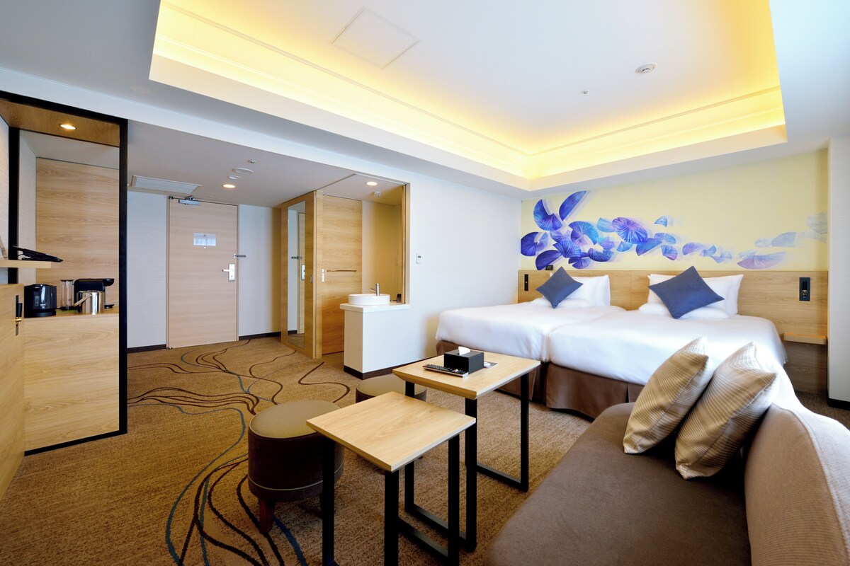 A hotel room with a bed, a sofa, a small table, a desk, a mirror, and a wall with a floral design.