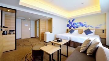 A hotel room with a bed, a sofa, a small table, a desk, a coffee machine, and a bathroom visible through an open door.