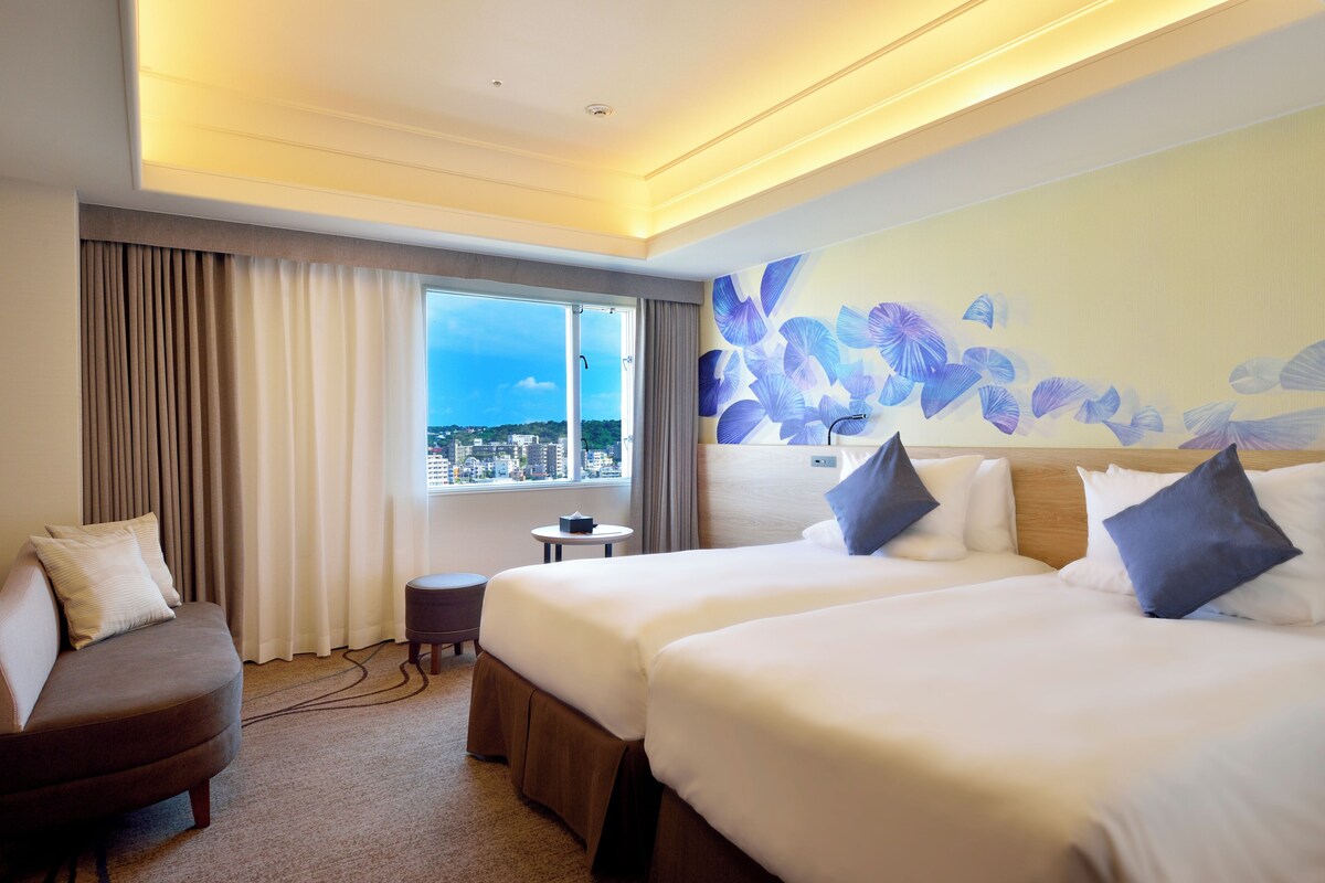 A hotel room with two beds, a sofa, a small table, and a view of the city through large windows.
