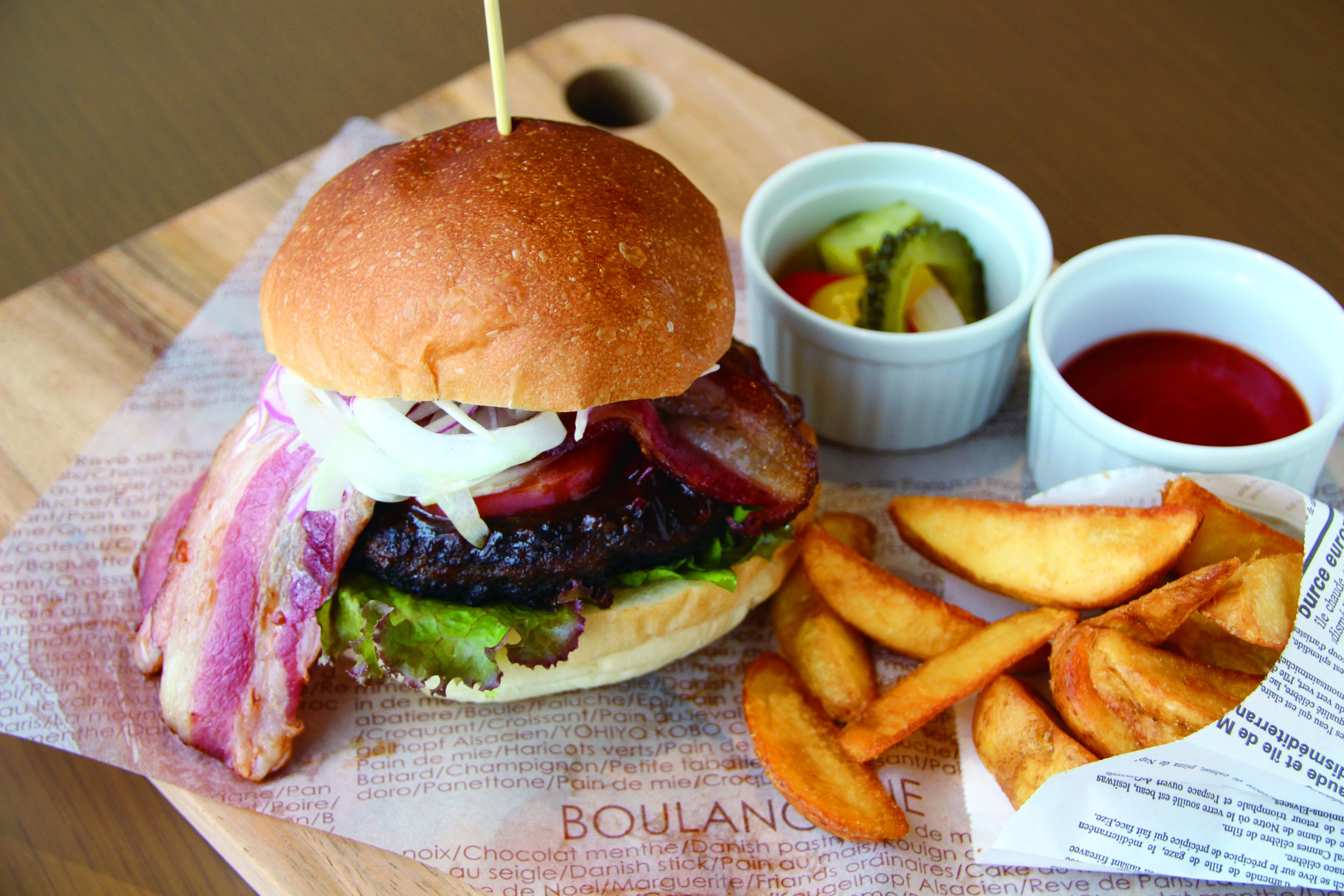 A burger with bacon, lettuce, and a bun, accompanied by fries and two dipping sauces on a wooden board.