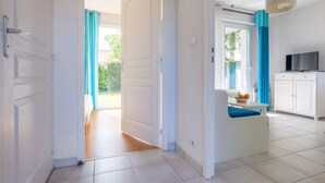 Ein helles Zimmer mit einer weißen Tür, einer kleinen Sitzecke mit einem blauen Kissen, einem Fernseher und einem Fenster mit blauen Vorhängen.