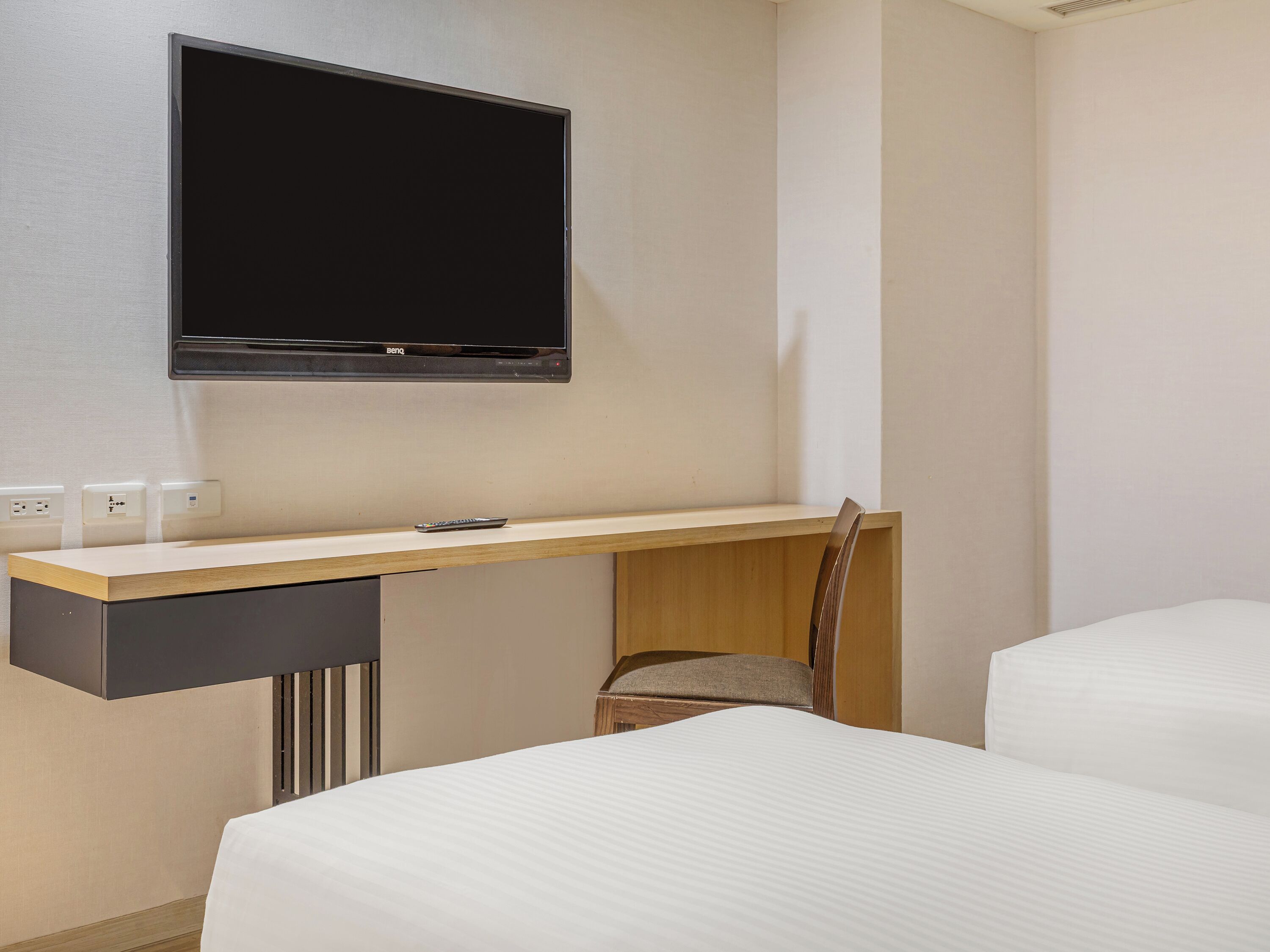 Ein Hotelzimmer mit einem Bett, einem Schreibtisch mit Stuhl, einem an der Wand befestigten Fernseher und einer Fernbedienung auf dem Schreibtisch.