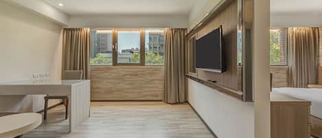 A modern hotel room with a flat-screen TV mounted on the wall, a dining table, a sofa, and a large window offering a city view.