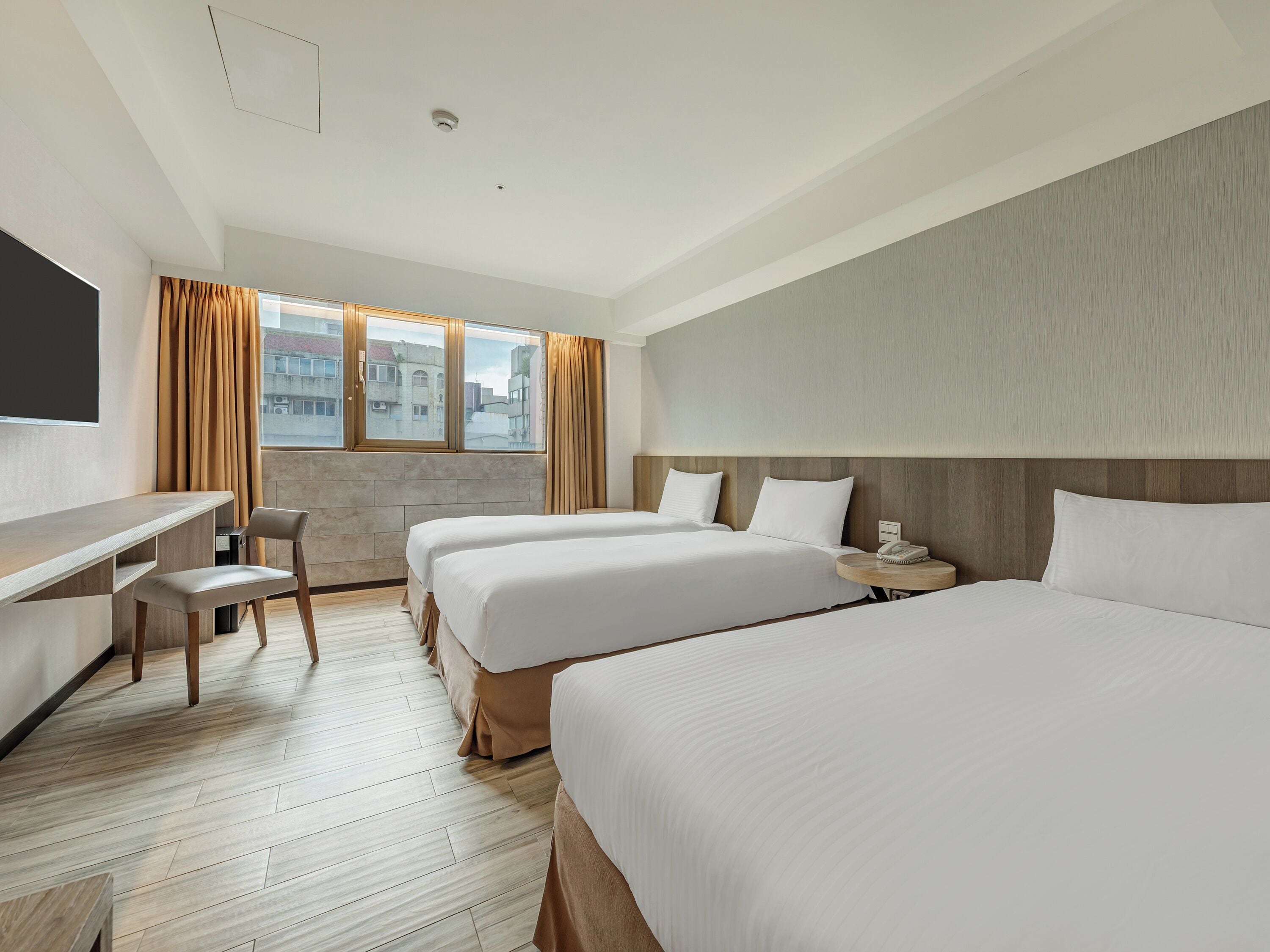 A hotel room with two beds, a desk, a chair, a bedside table with a lamp, and a large window with curtains.