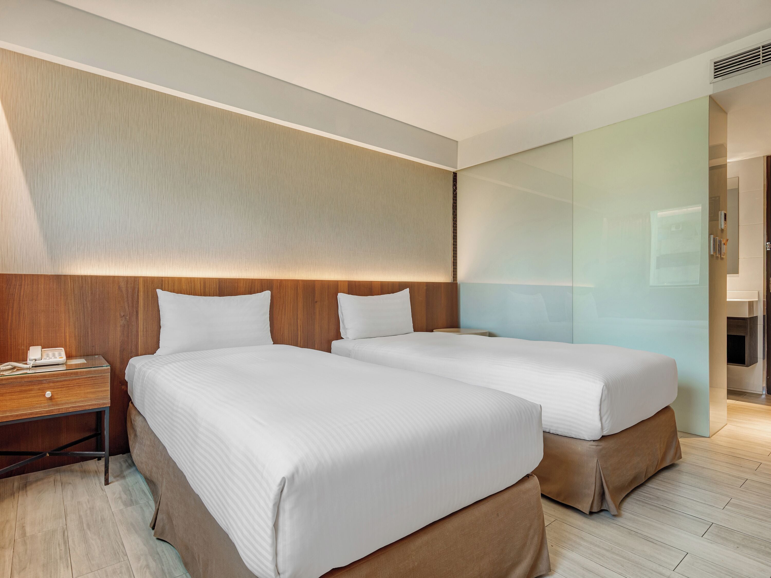 Une chambre d’hôtel avec deux lits, une tête de lit en bois, une table de chevet avec un téléphone et une cloison en verre.