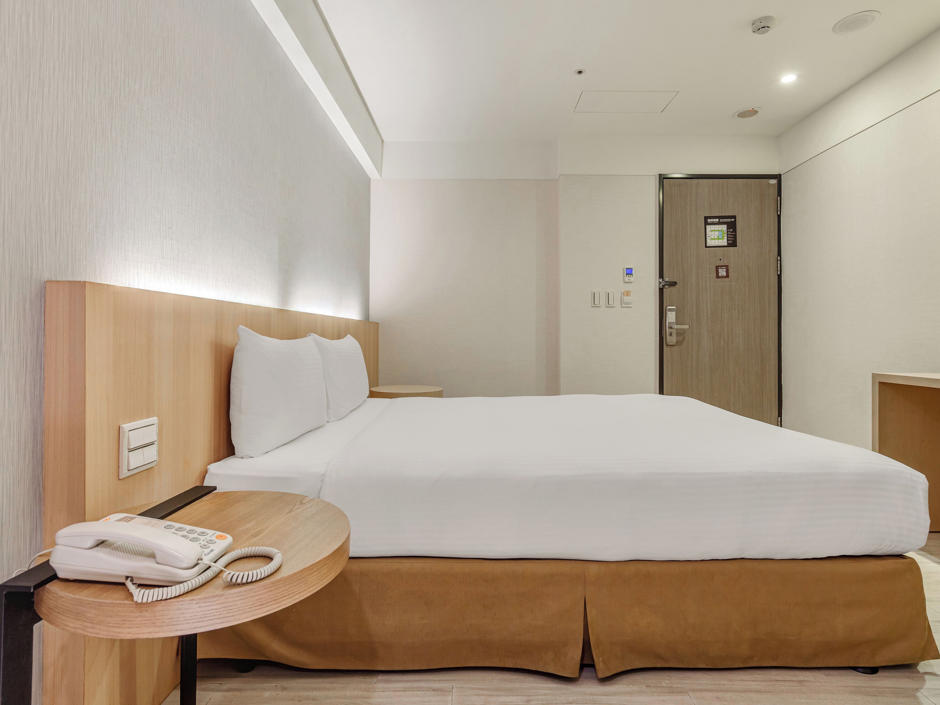 A hotel room with a bed, bedside table with a phone, a door with a lock, and a wall-mounted light switch.