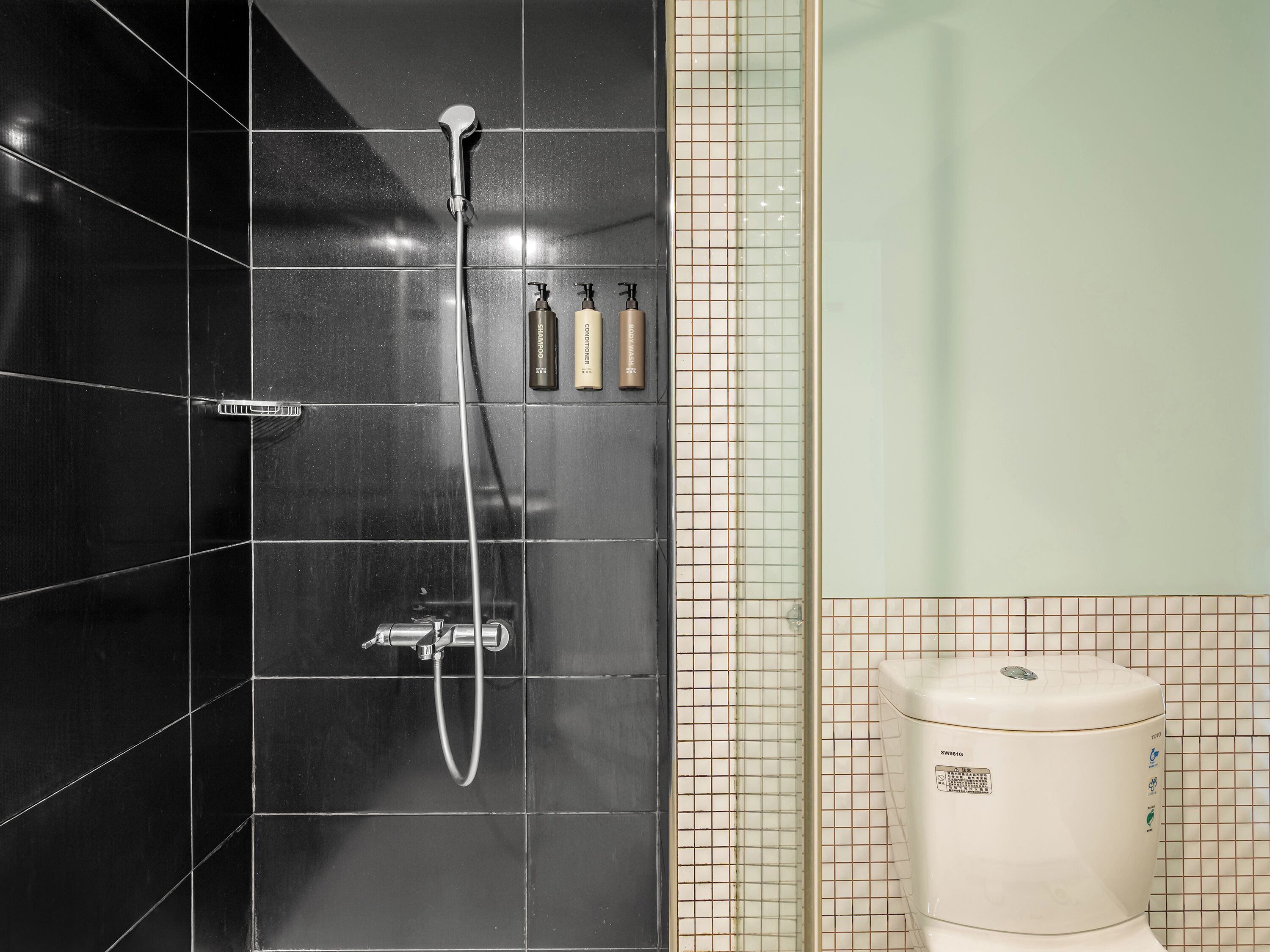 Une salle de bain avec des carreaux noirs, une douche équipée d’un pommeau de douche à main et des toilettes portant un label indiquant qu’elles permettent d’économiser l’eau.