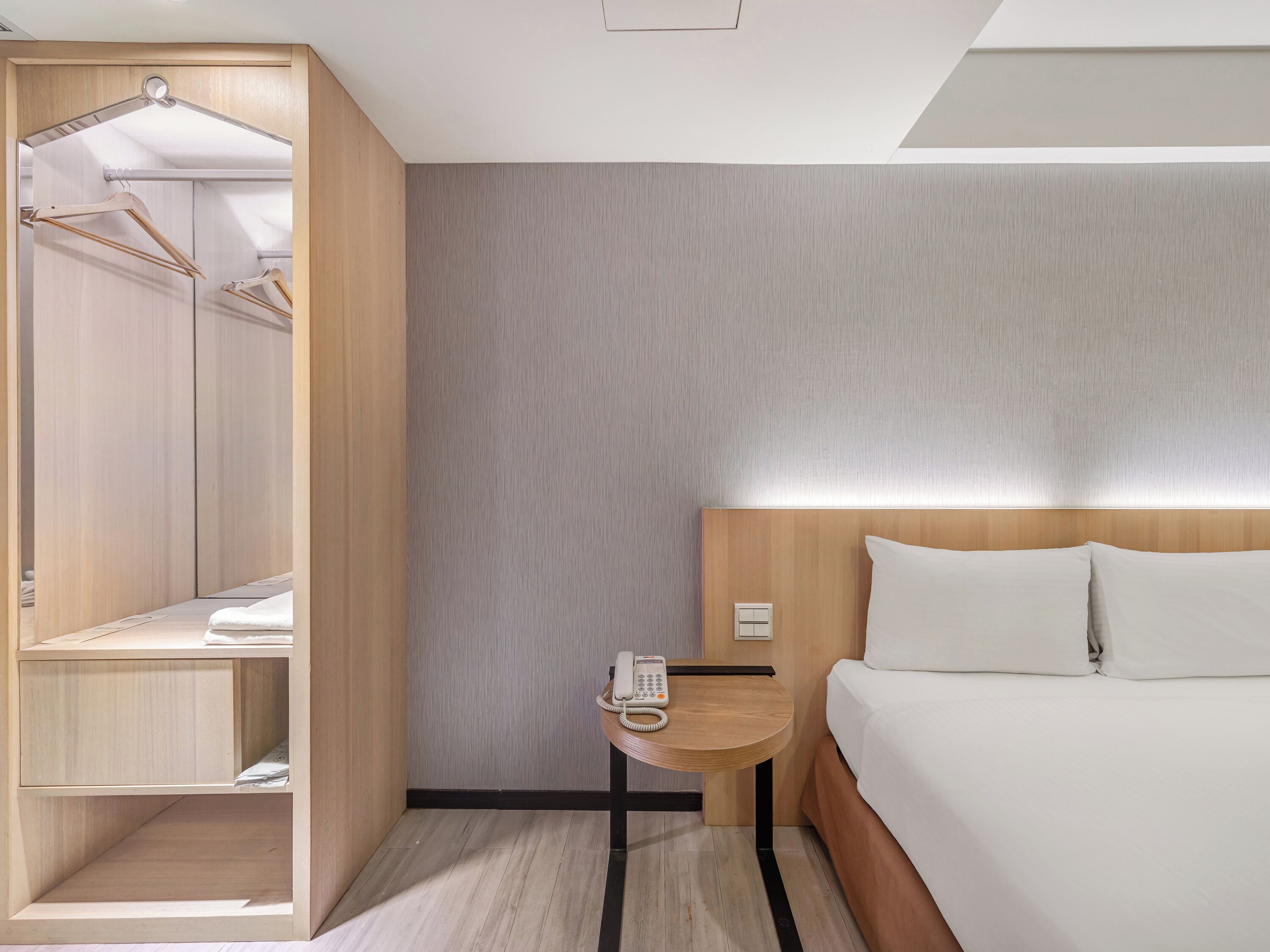 A hotel room with a bed, a bedside table with a phone, a wooden wardrobe, and a mirror.