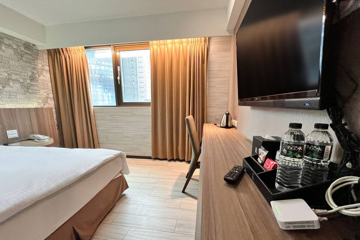 A hotel room with a bed, a desk with a computer, a TV, and a window with curtains.