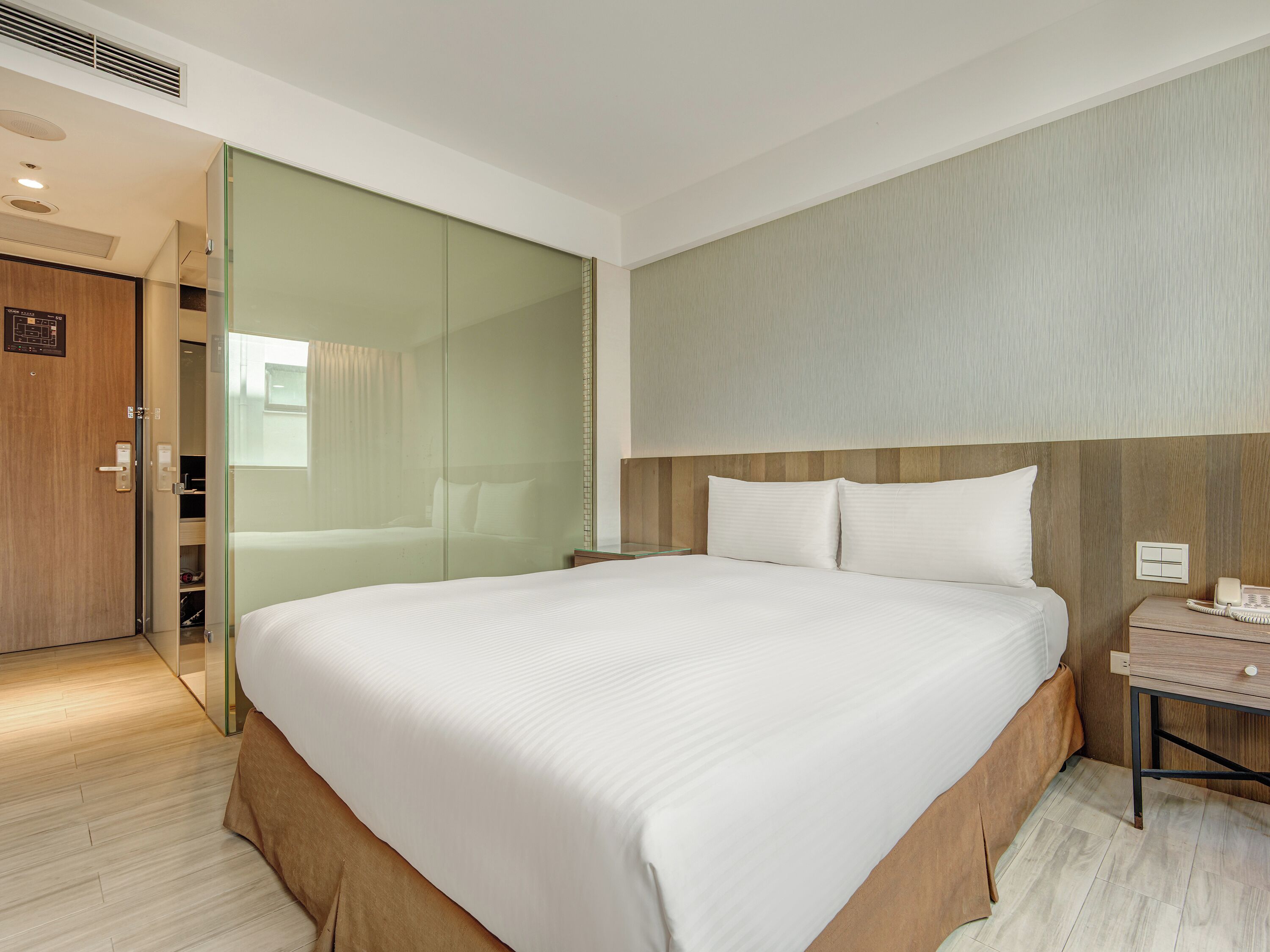 A hotel room with a large bed, a bedside table with a lamp, a wooden wardrobe, and a glass partition.