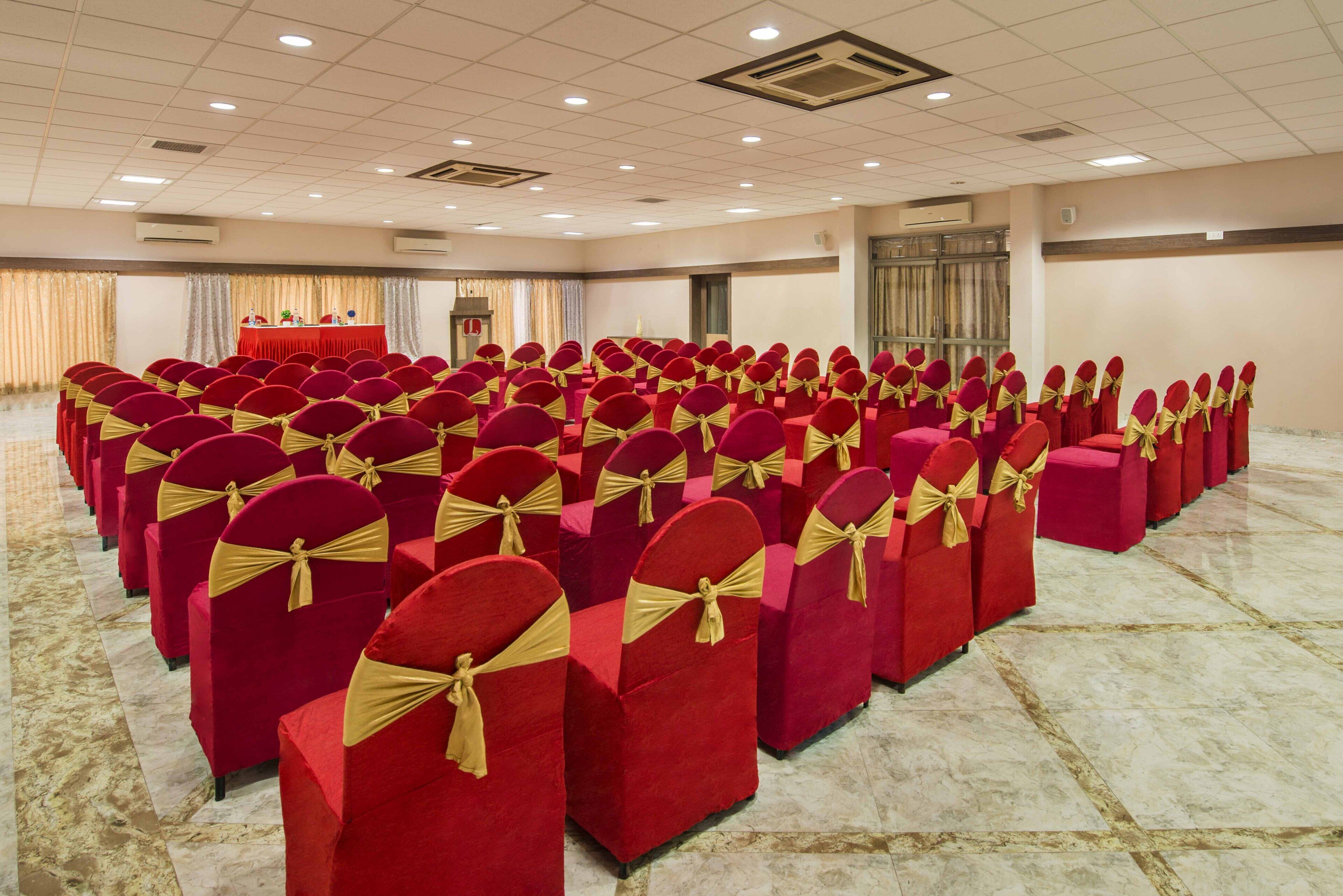 Un salón de conferencias con filas de sillas rojas con cintas doradas, un atril y una mesa con mantel rojo.