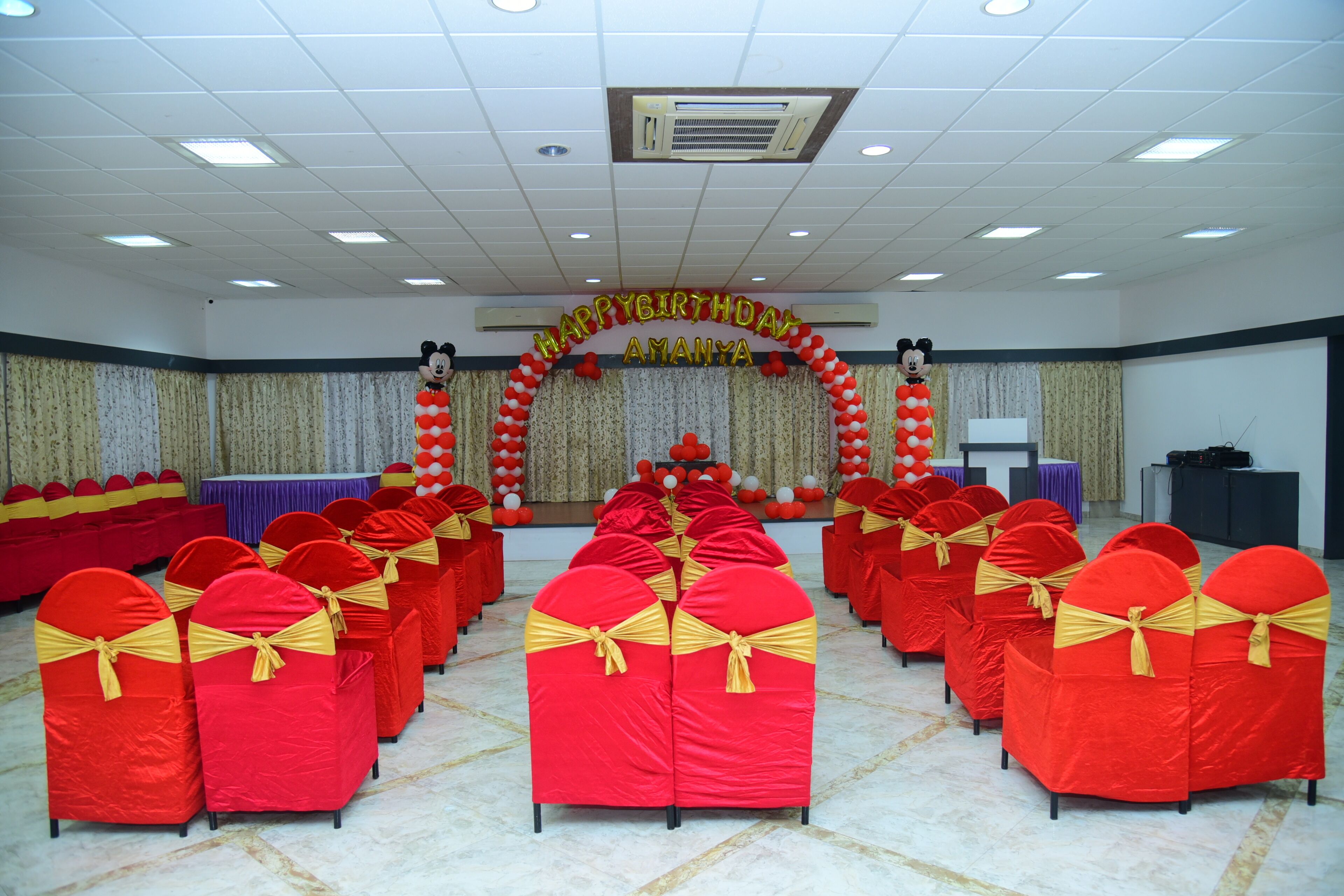 A conference room with rows of red chairs with yellow sashes, a stage with a 'Happy Birthday Amanya' banner, and a backdrop with balloons.