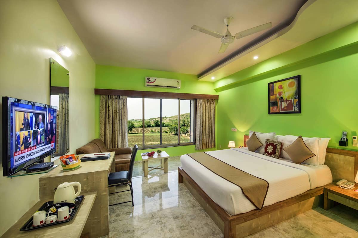 Quarto de hotel com cama, escrivaninha com televisão, cadeira, janela com cortinas, ventilador de teto e um quadro na parede.