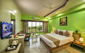 A hotel room with a bed, a desk with a TV, a chair, a window with curtains, a ceiling fan, and a painting on the wall.