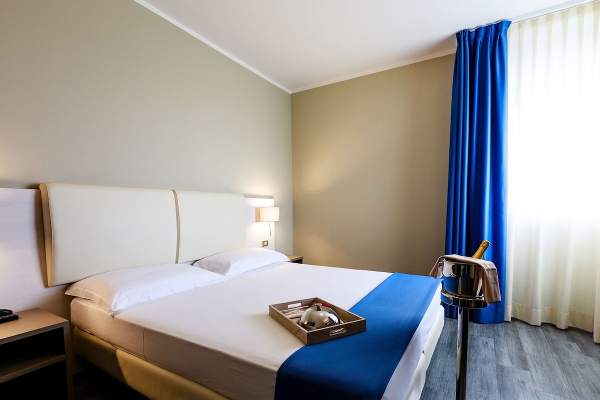 Habitación de hotel con cama, mesita de noche, lámpara, minibar y ventana con cortinas.