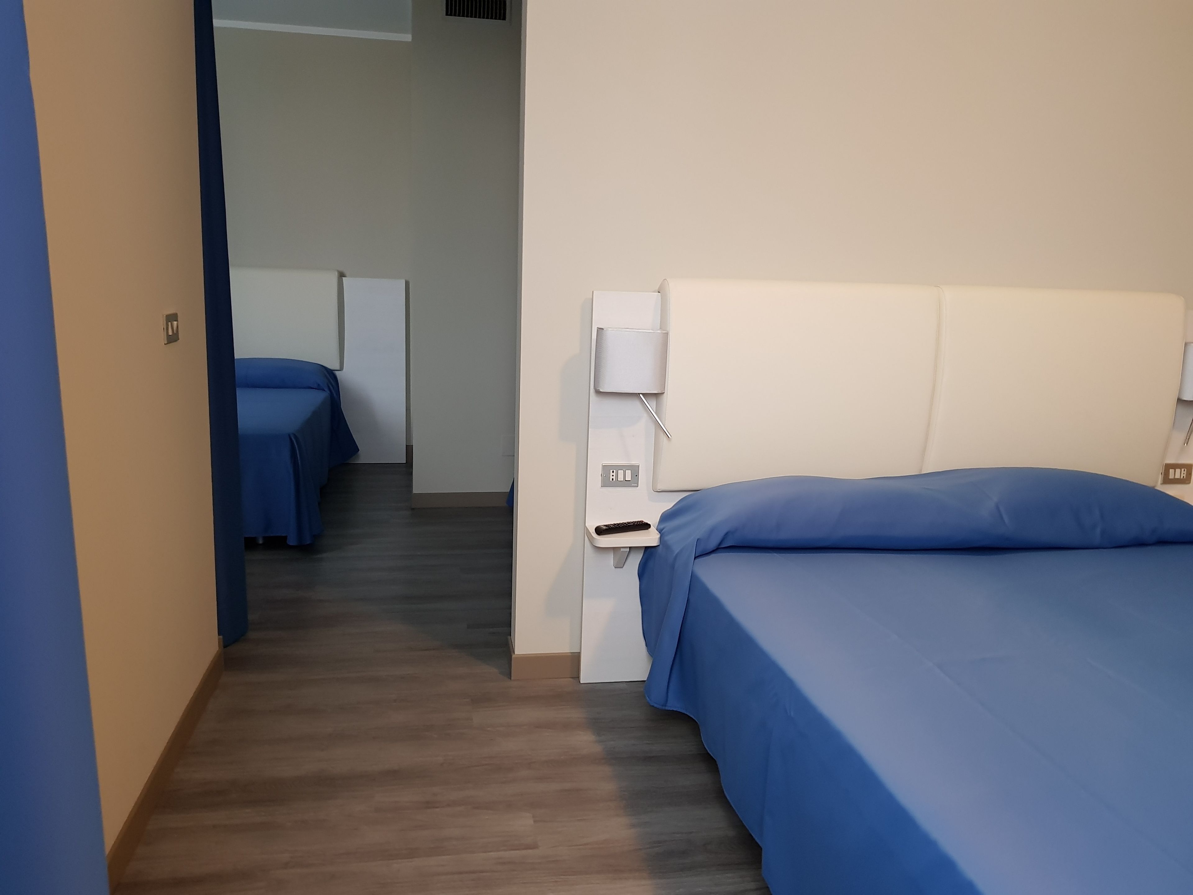 Egy szállodai szoba, amelyben egy egyszemélyes ágy, egy éjjeliszemély és egy falra szerelt lámpa található.