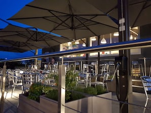 An outdoor dining area with multiple tables and chairs, umbrellas, and a modern architectural design. - Hotel Oleggio Malpensa (Oleggio)