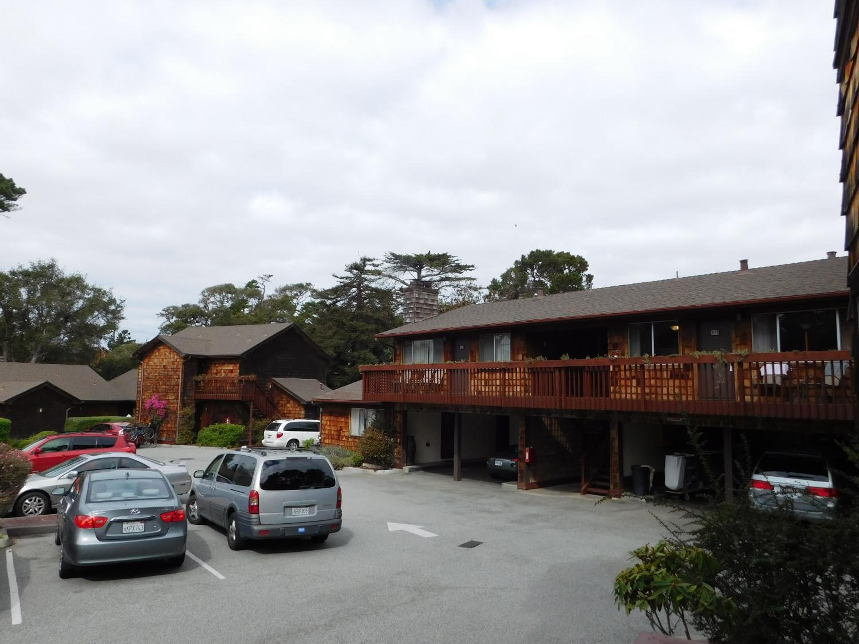 A parking lot with several cars, a building with a wooden balcony, and a brick structure in the background.