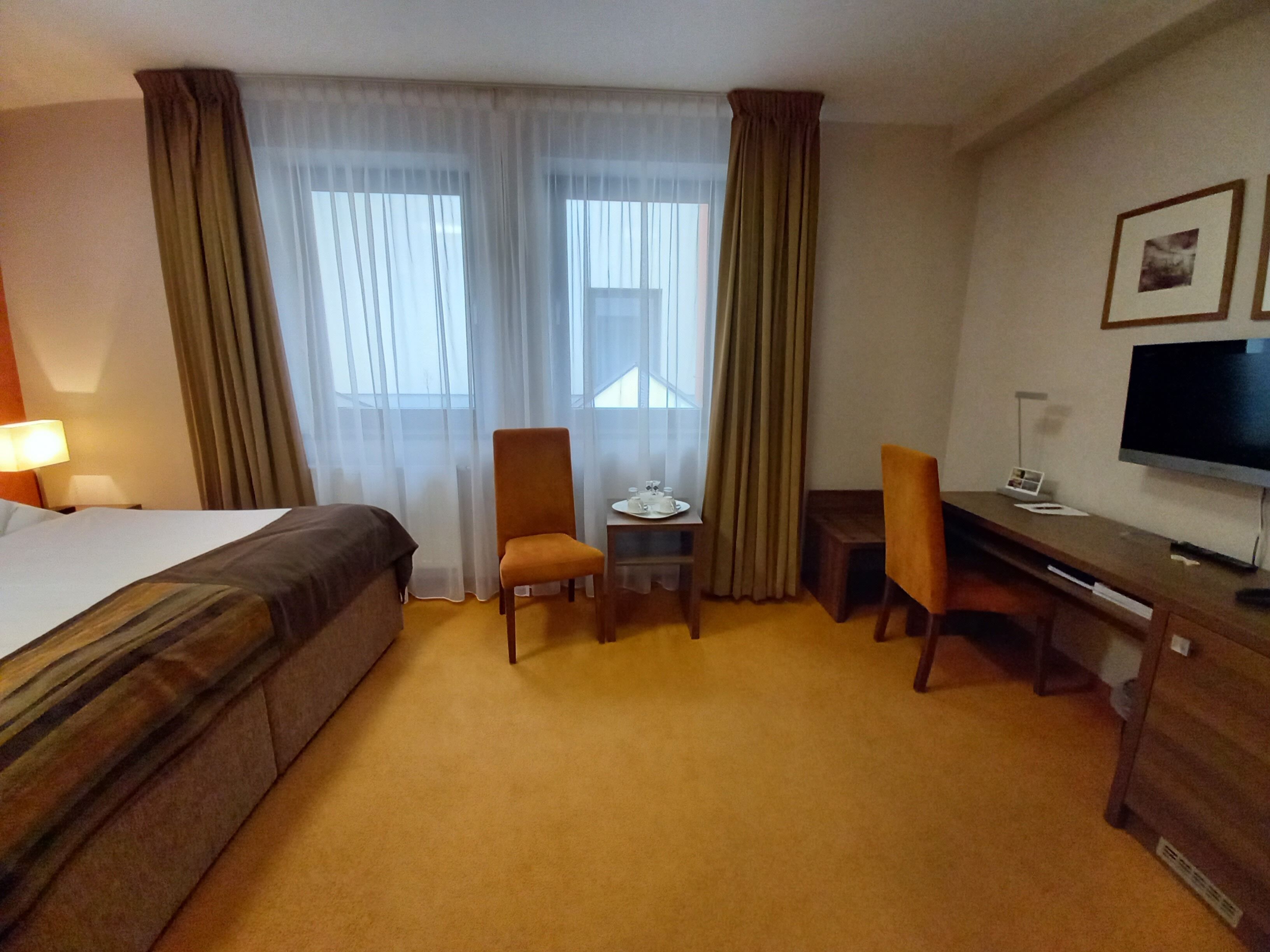 Una habitación de hotel con una cama, dos sillones, un escritorio, un televisor y una ventana con cortinas.