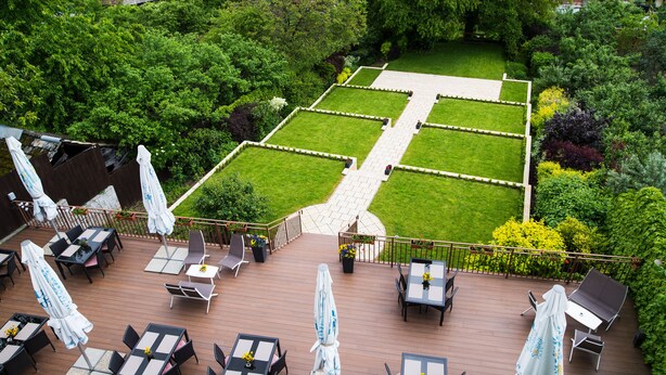 Eine Terrassengestaltung im Freien mit verschiedenen Sitzbereichen, darunter Tische und Stühle, umgeben von üppiger Bepflanzung und einem gepflegten Rasen.