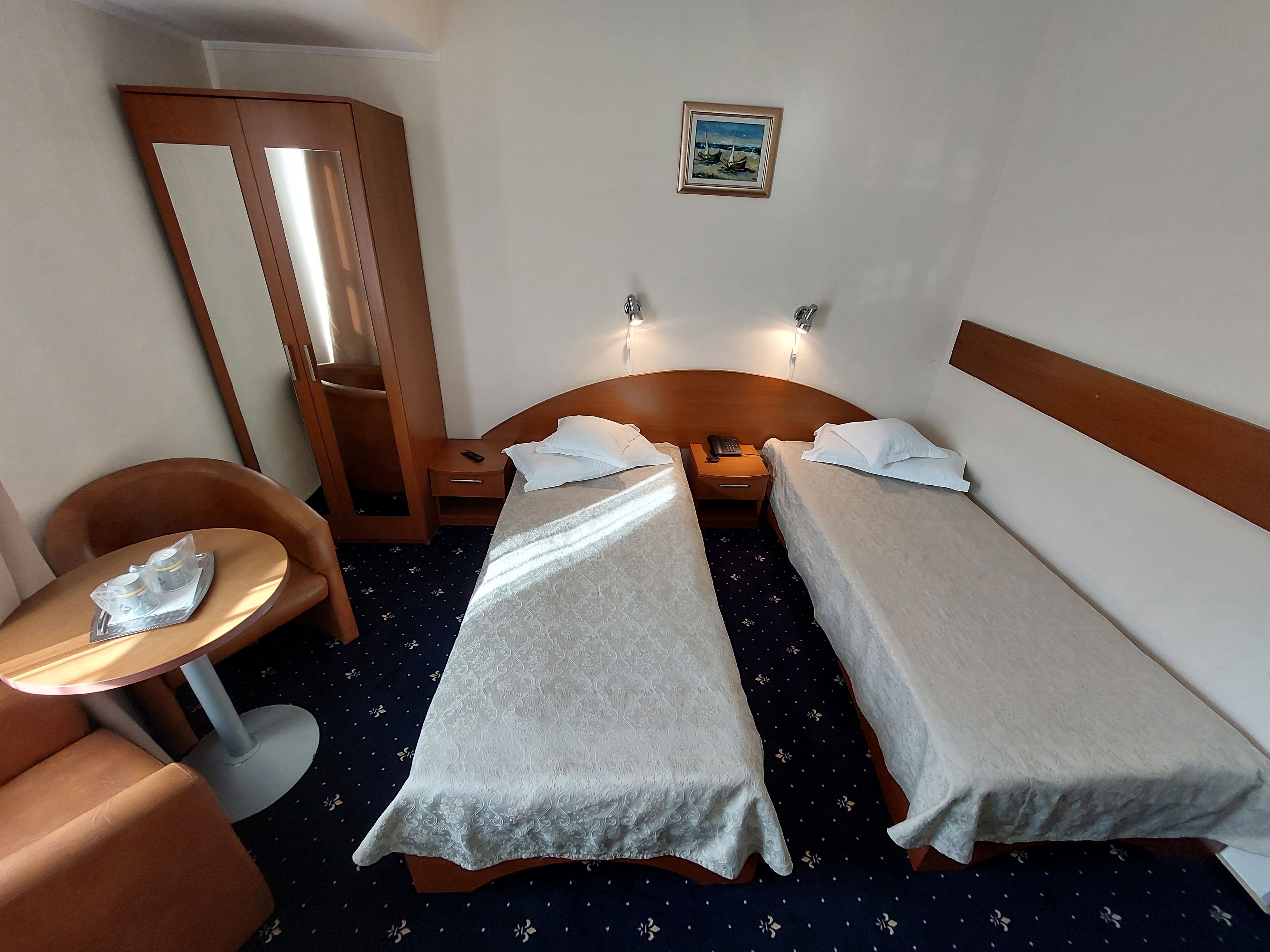 A hotel room with two single beds, a small table, a chair, a bedside table with a lamp, and a mirror.