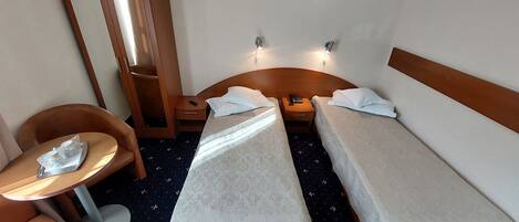 A hotel room with two single beds, a small table, a chair, a bedside table with a lamp, and a mirror.