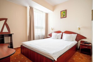 A hotel room with a bed, a desk, a chair, a nightstand, a lamp, and a painting on the wall.