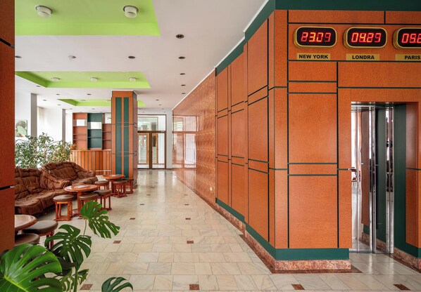 A hotel lobby with a modern design, featuring a seating area with sofas and chairs, a reception desk, and a digital clock displaying times for New York, London, and Paris. - Egreta (Tulcea)