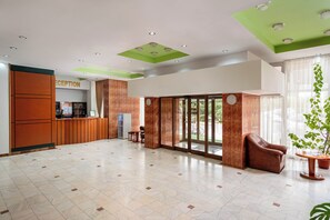 A hotel reception area with a reception desk, a seating area with a sofa and chairs, and a large glass door leading outside.