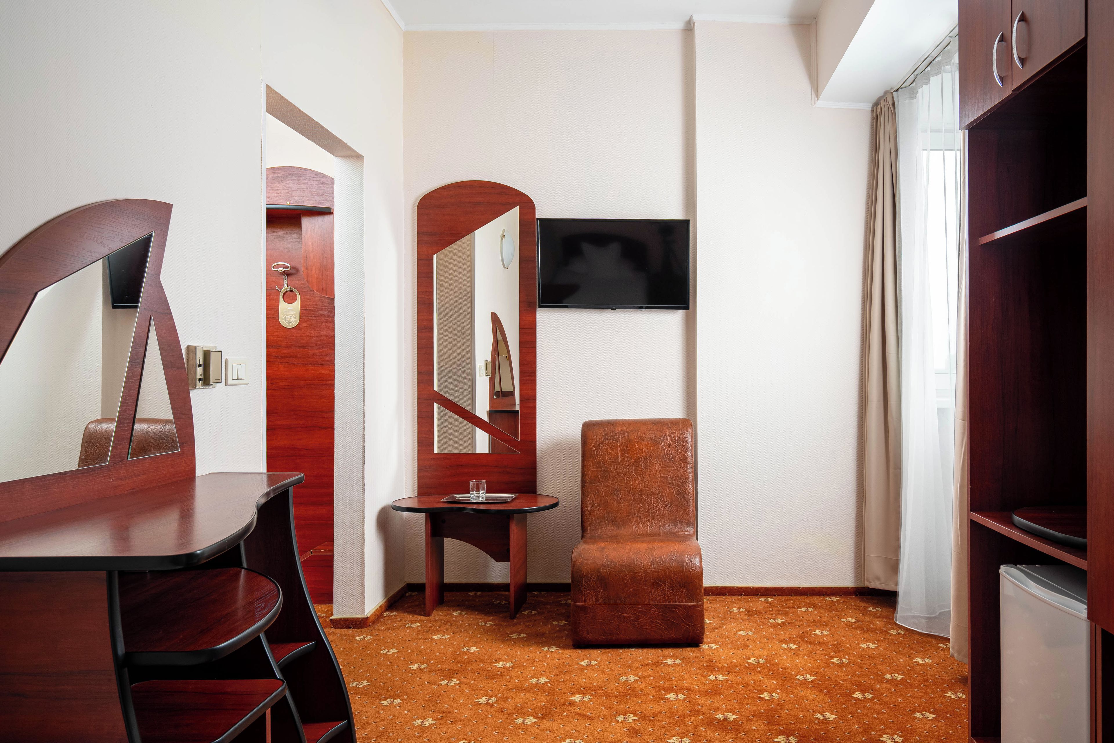 A hotel room with a wooden desk, a small side table, a single chair, a television mounted on the wall, and a wardrobe.