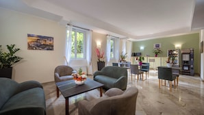 Un salon moderne comprenant un canapé, des fauteuils, une table basse et un coin repas avec une table et des chaises. On y trouve un tableau au mur, une fenêtre avec des rideaux et un distributeur automatique.