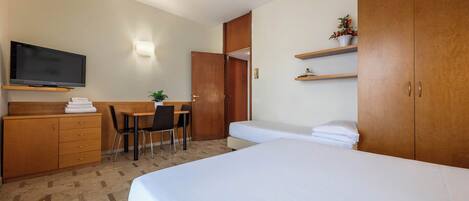 A hotel room with a bed, a desk with a chair, a television, a wardrobe, and a shelf with a plant.