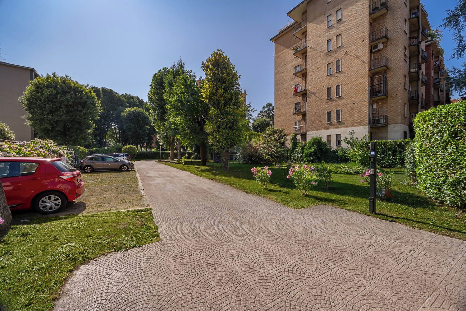A residential area with a paved walkway, parked cars, and a multi-story apartment building.