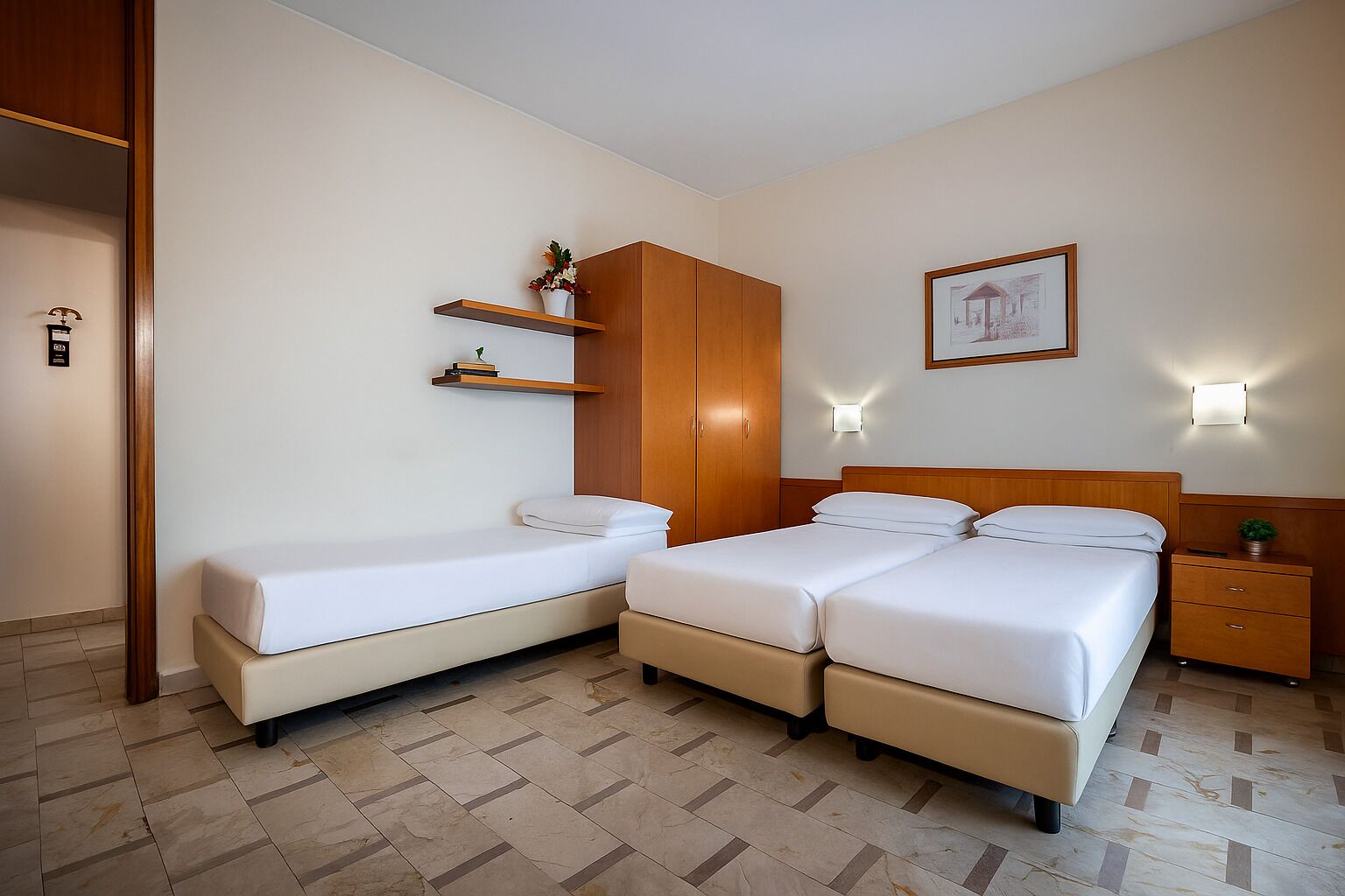 Habitación de hotel con cama doble, cama individual, armario de madera, mesitas de noche y un cuadro enmarcado en la pared.