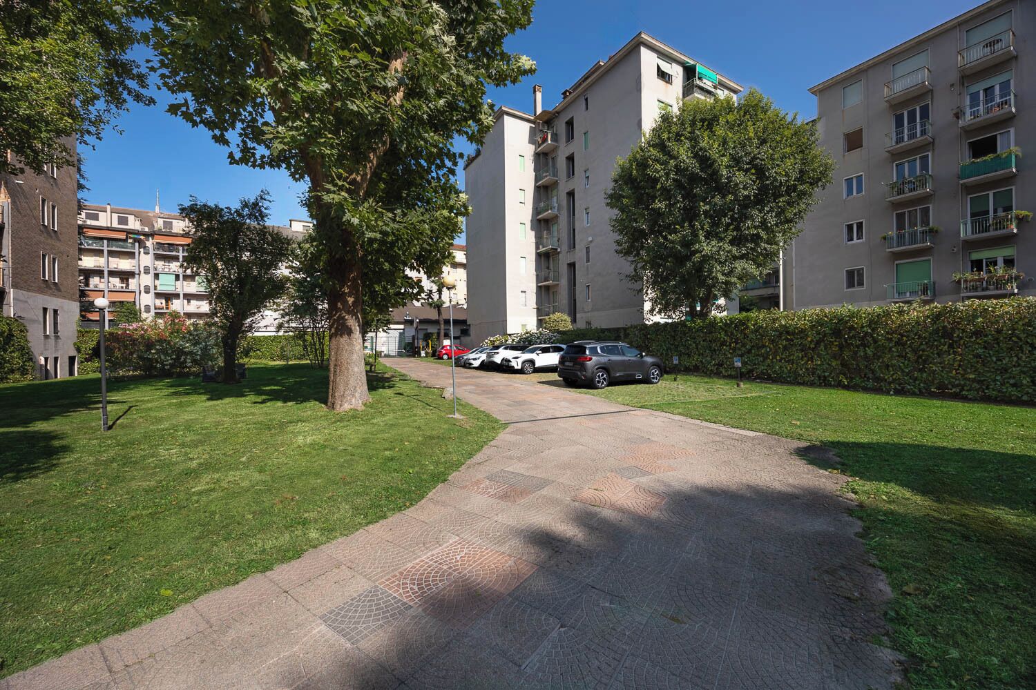 A residential building with a paved pathway, parked cars, and surrounding greenery.