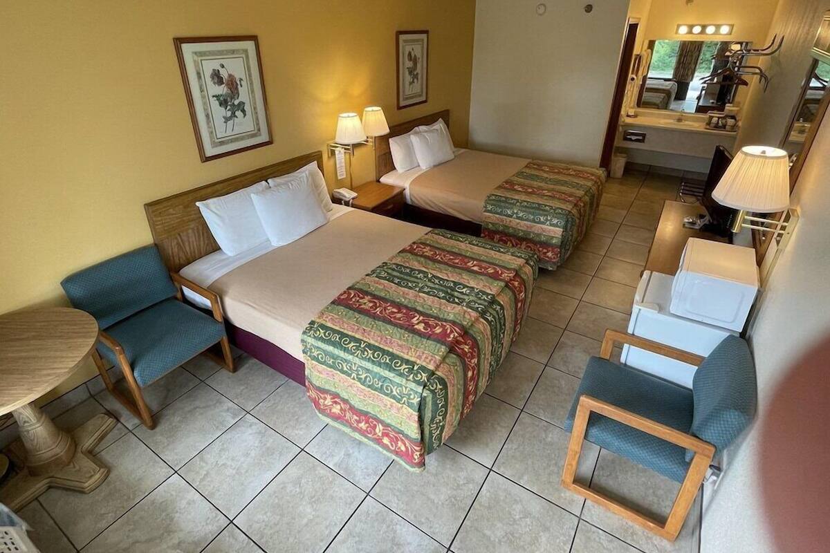Habitación de hotel con dos camas, una mesita, una silla, un escritorio y un baño con espejo y lavamanos.