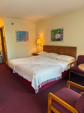 Habitación de hotel con cama, mesita de noche, lámpara, una silla y dos obras de arte en la pared.