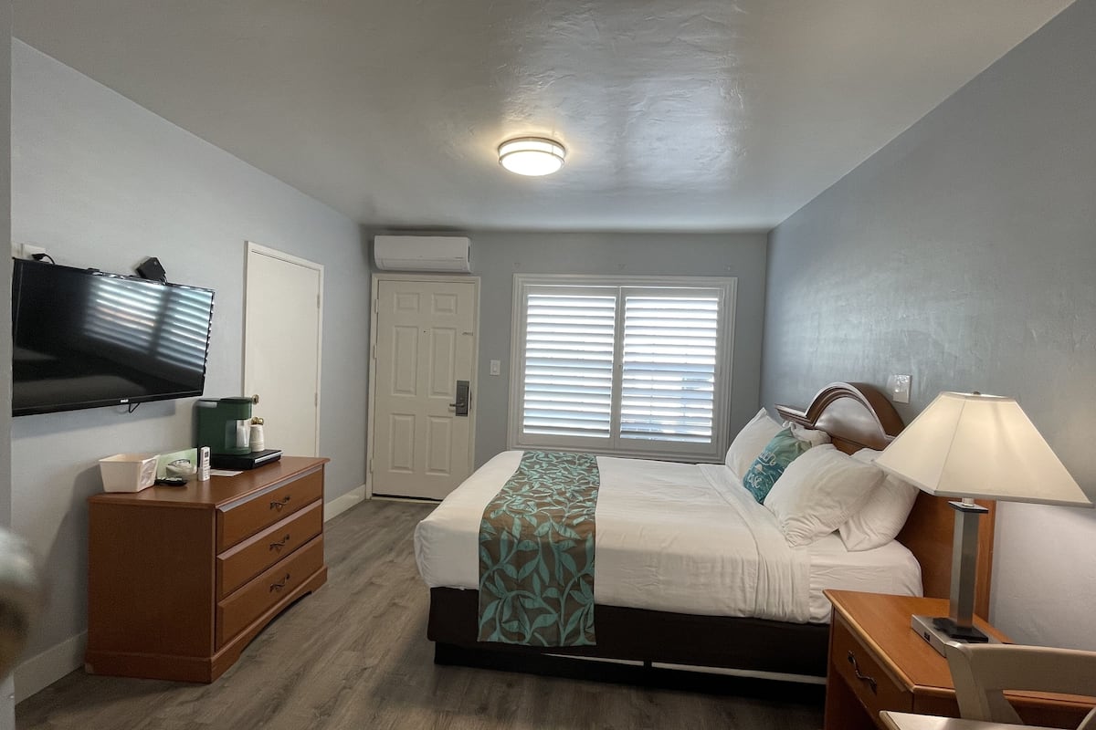 A bedroom with a bed, dresser, TV, and a window with blinds.